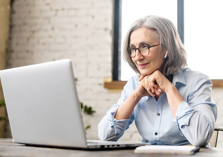 Een vrouw met grijs haar en een bril zit voor een laptop. Met een oordopje in. Ze kijkt vriendelijk.