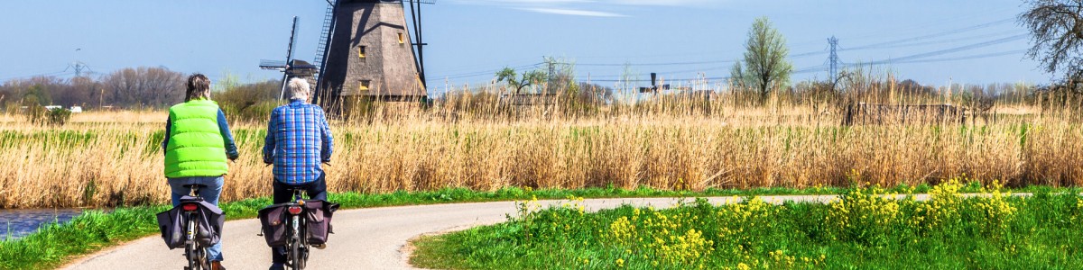 Twee mensen fietsen over een polderweg. Achterin zie je een molen en een korenveld en blauwe lucht.