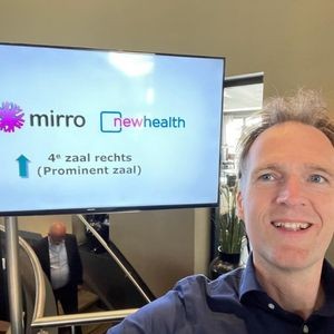 Foto van directeur Frank. Hij staat voor een scherm met Mirro en NewHealth erop. Hij lacht breed.