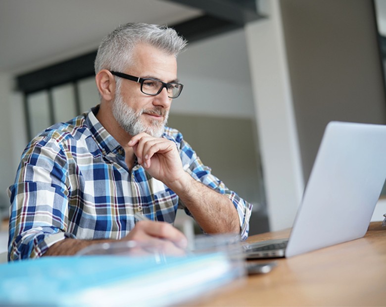 Een man met grijs haar en een bril gebruikt een laptop. Hij houdt een pen vast en glimlacht licht.